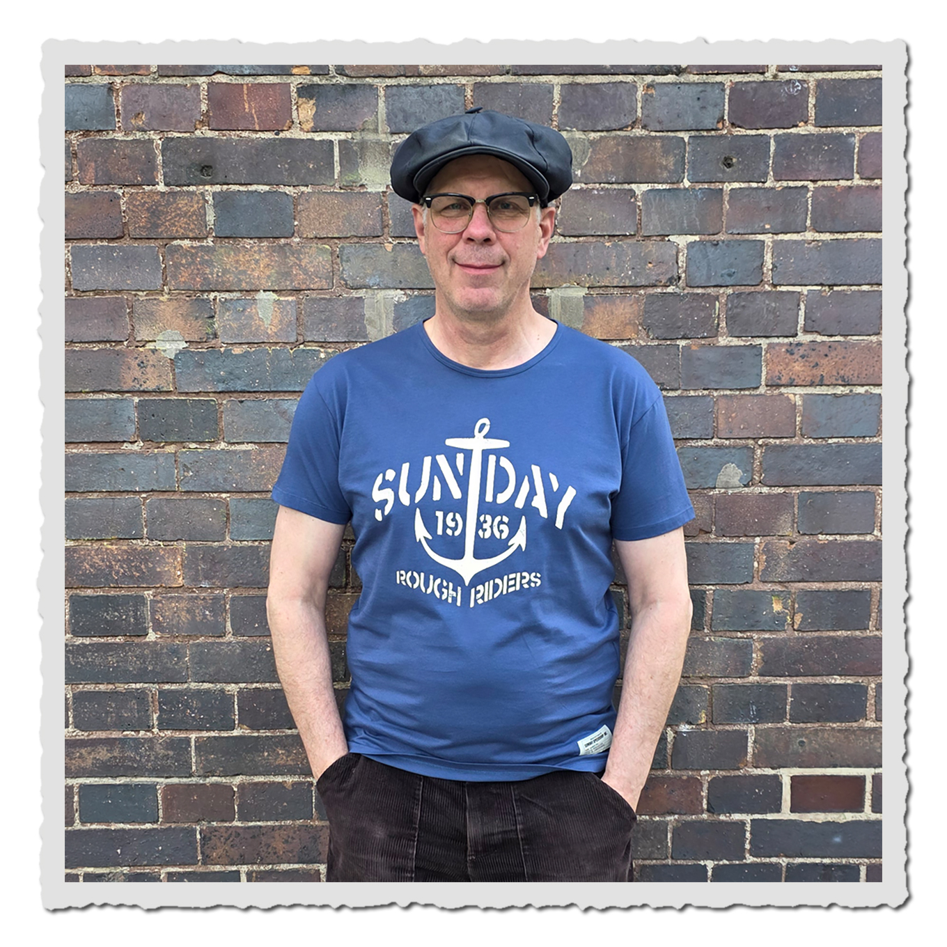 Mann trägt blaues T-Shirt mit hellem Ankerdruck vor einer Backsteinwand, Vorderansicht.